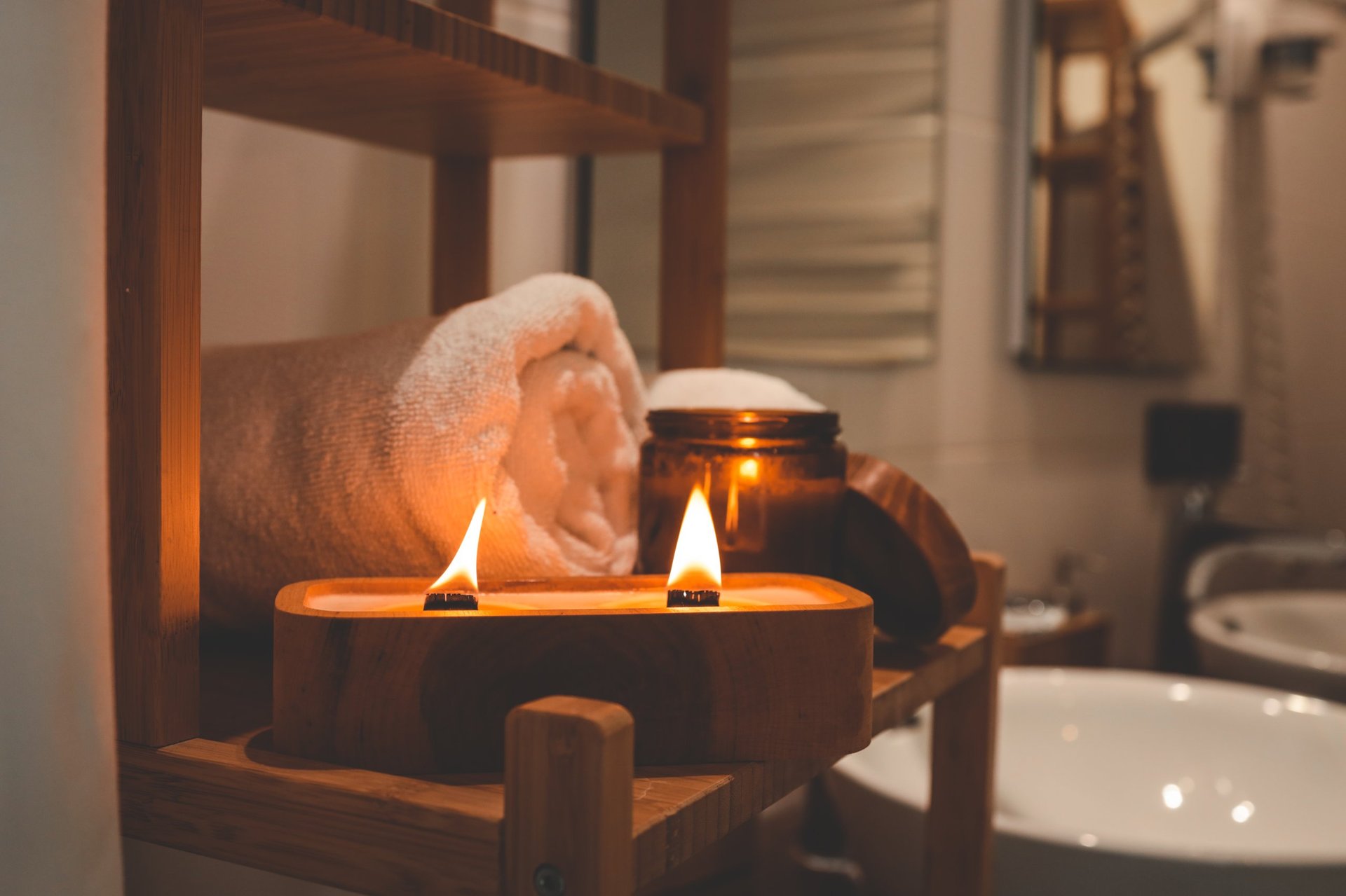 Burning wooden wick candle with amber jar and rolled towel on bamboo shelf in cozy bathroom, warm spa ambiance